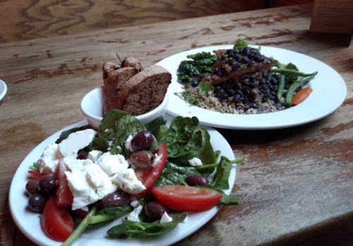 Two plates of food on a wooden table: one with salad and feta, the other with quinoa, black beans, and greens.