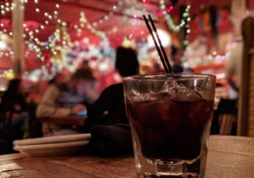 A glass of dark drink with ice sits on a table, with a blurred, festive background of lights and people.