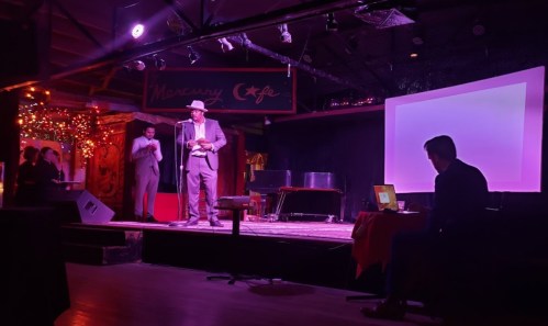 A performer in a hat stands on stage at a dimly lit venue, with a piano and audience in the background.