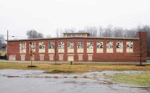 Abandoned brick building with large, broken windows, surrounded by a grassy area and overcast sky.