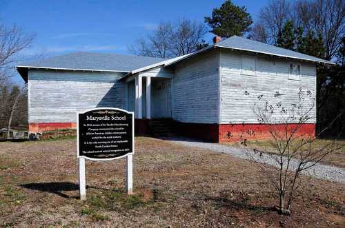 Historic Marysville School building with a sign in front, surrounded by trees and a gravel path.