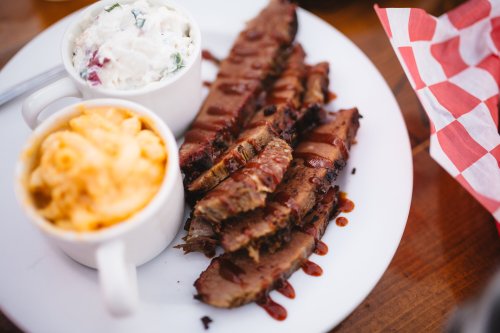 A plate featuring sliced brisket drizzled with sauce, alongside mac and cheese and a cup of potato salad.