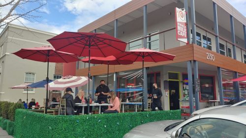 Outdoor café scene with red umbrellas, people dining, and a modern building in the background.