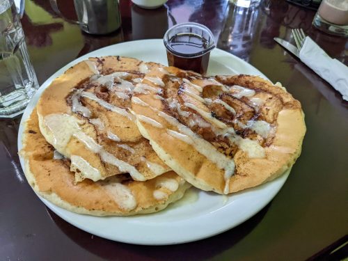 Three large cinnamon roll pancakes drizzled with icing, served with a small cup of syrup on a white plate.