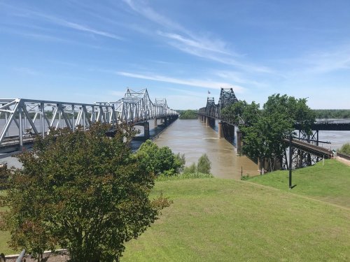 A view of two bridges spanning a wide river, surrounded by green trees and a clear blue sky.