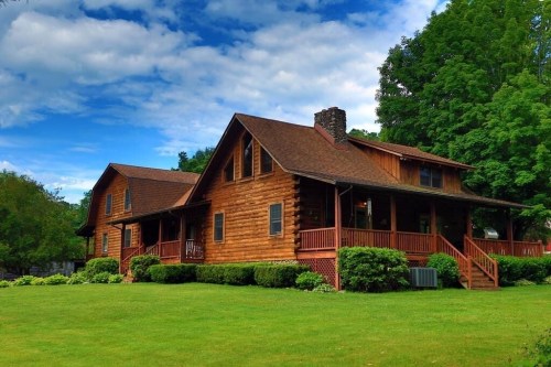 A charming log cabin home with a large lawn, surrounded by trees and under a blue sky with fluffy clouds.