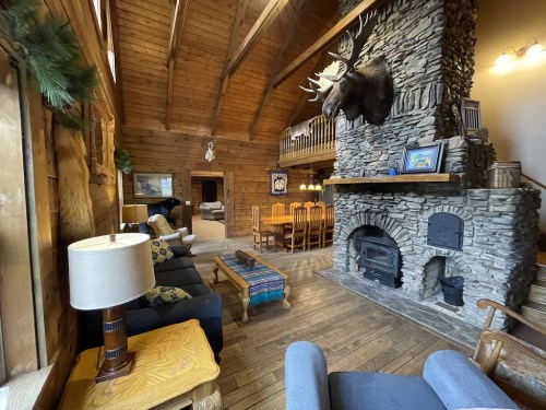 Cozy cabin interior with wooden walls, stone fireplace, moose head mount, and a dining area. Warm, rustic atmosphere.