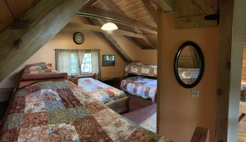 Cozy cabin interior with wooden beams, featuring multiple beds and a mirror, illuminated by natural light.
