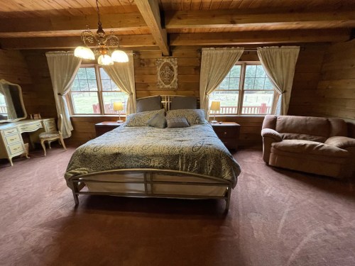 Cozy wooden bedroom with a large bed, two windows, a dresser, and a comfortable chair. Warm, inviting atmosphere.