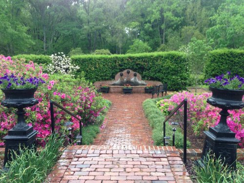 A brick pathway leads to a garden with vibrant flowers, benches, and a stone wall surrounded by lush greenery.