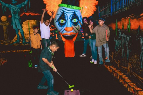 A group of people plays mini-golf in a glow-in-the-dark setting with a large clown figure in the background.