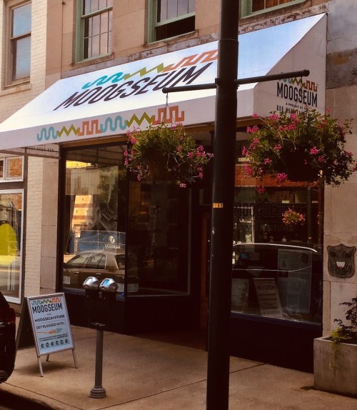 Exterior of the Moogseum with a colorful awning, flower baskets, and a sign outside on a sunny day.