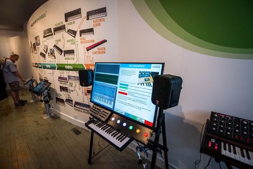 A display of vintage synthesizers and music technology, featuring screens and equipment in a museum setting.