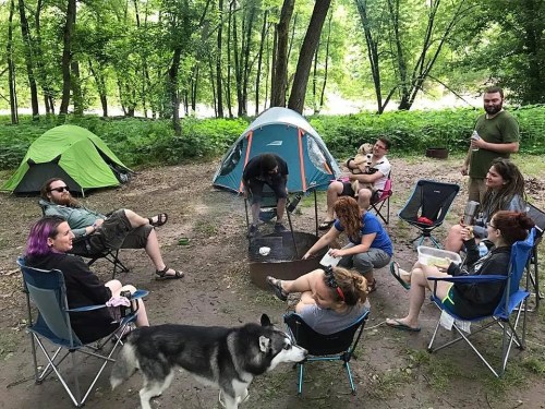 A group of people camping in a forest, sitting around a fire pit with tents and a dog nearby.