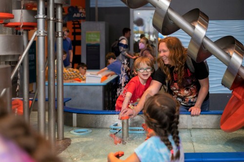 A child and an adult play with water at a hands-on exhibit, surrounded by other children exploring nearby activities.