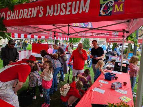 A busy outdoor event at a children's museum, with families engaging in crafts under a red tent.