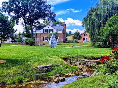 A scenic view of a house surrounded by greenery, with a sculpture and a small stream in the foreground.