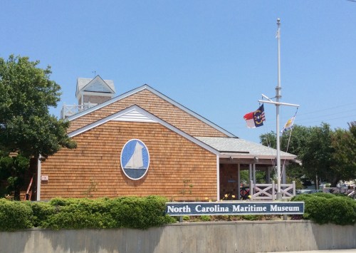 Exterior of the North Carolina Maritime Museum, featuring a wooden building and flags, surrounded by greenery.