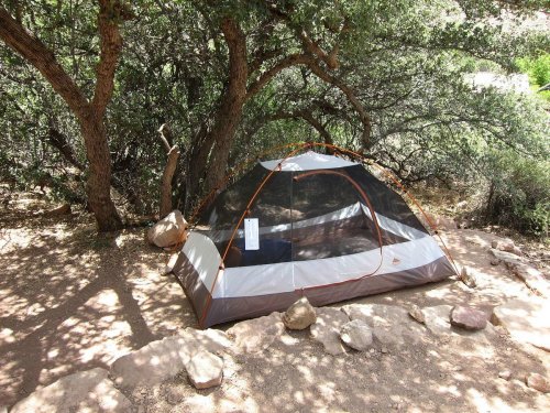 A camping tent set up under trees, surrounded by rocks and dirt in a natural outdoor setting.