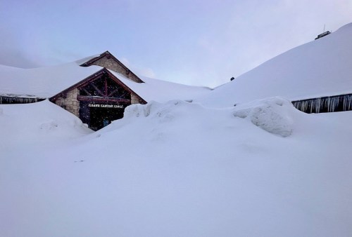 A snow-covered building with a peaked roof, surrounded by deep snowdrifts under a cloudy sky.
