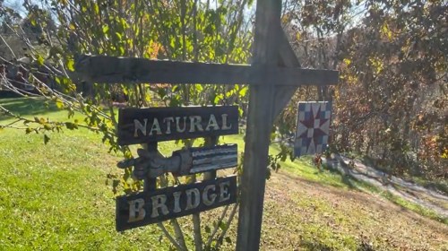 A wooden sign reading "Natural Bridge" with a decorative quilt pattern in the background, surrounded by greenery.