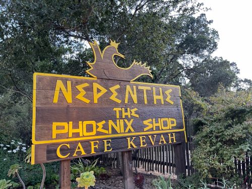 Wooden sign for Nepenthe featuring "The Phoenix Shop" and "Cafe Kevah," surrounded by greenery.