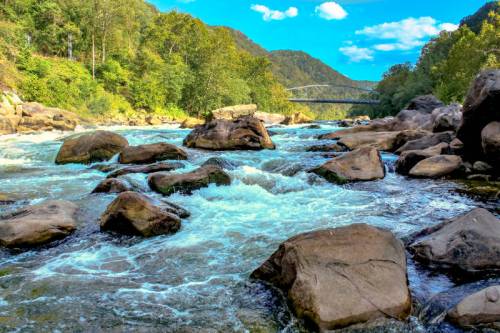 A serene river flows over rocks, surrounded by lush greenery and mountains under a bright blue sky with fluffy clouds.