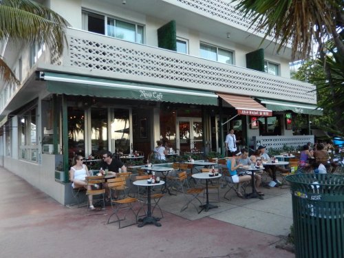 Outdoor café scene with patrons dining at tables, surrounded by palm trees and a bustling street atmosphere.