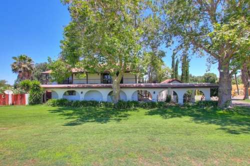 A spacious two-story house surrounded by lush greenery and large trees on a sunny day.
