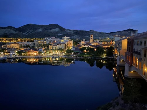A serene lakeside view at dusk, with illuminated buildings reflecting on the water and mountains in the background.