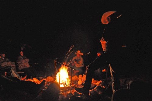 A group of people sitting around a campfire at night, with one person playing guitar and others enjoying the warmth.