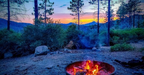 A campfire burns in a metal ring, surrounded by trees, with a colorful sunset in the background.