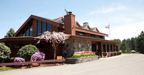 A modern wooden building with large windows, surrounded by colorful flowers and trees, under a clear blue sky.