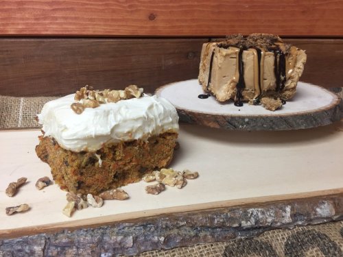 A slice of carrot cake with cream cheese frosting beside a round cheesecake drizzled with chocolate sauce and topped with nuts.