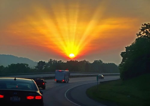 A vibrant sunset with rays shining over a highway, framed by trees and silhouettes of cars.