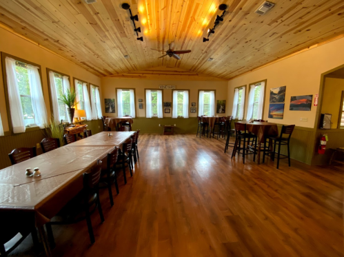 A spacious room with wooden floors, large windows, and tables set for dining, featuring a cozy, inviting atmosphere.