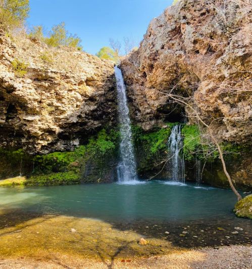 A serene waterfall cascades into a clear pool, surrounded by rocky cliffs and lush greenery under a blue sky.