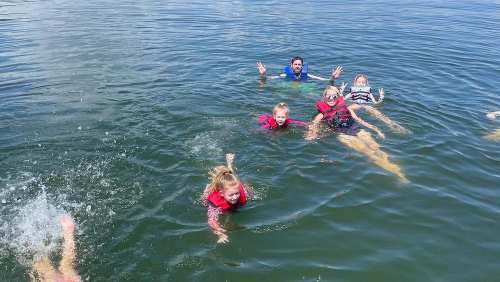 A group of children and an adult swim in a lake, all wearing life jackets and smiling, enjoying a sunny day.