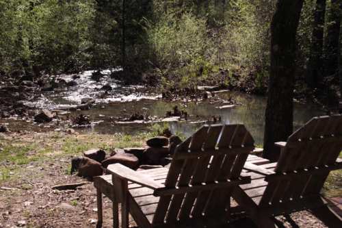 Two wooden chairs by a serene stream, surrounded by lush greenery and sunlight filtering through the trees.