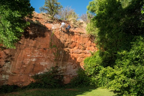 Two climbers ascend a rocky cliff surrounded by lush greenery and blue sky.