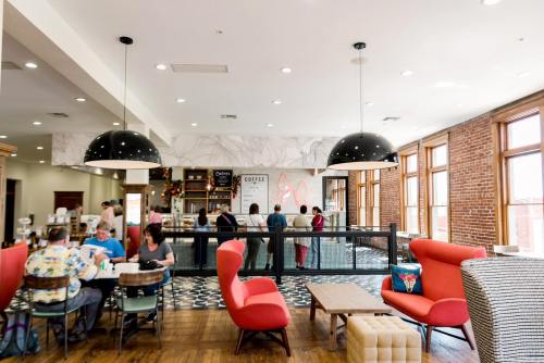 A cozy café interior with red chairs, wooden tables, and patrons enjoying coffee at the counter under large windows.