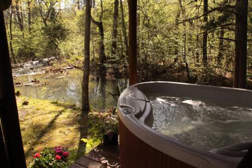A hot tub on a wooden deck overlooks a serene forest and a gently flowing stream.