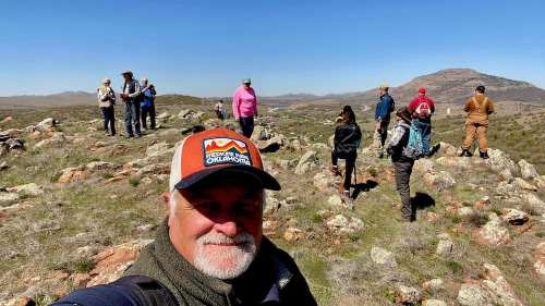 A group of people exploring a rocky landscape with hills in the background on a clear day. A man takes a selfie in the foreground.