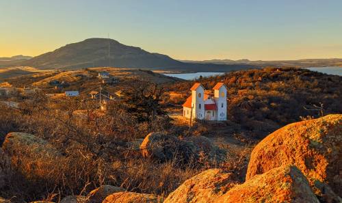 A scenic view of a white church with orange roofs, surrounded by rocky terrain and hills at sunset.