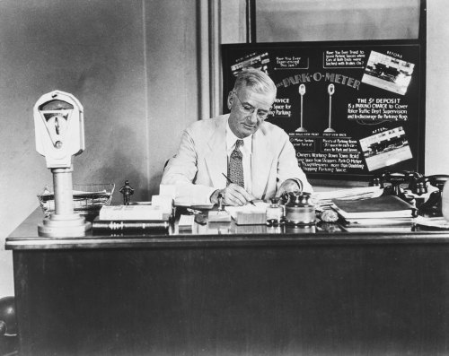 A man in a suit sits at a desk, writing, with a parking meter and informational poster in the background.
