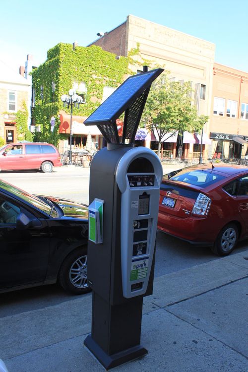 A solar-powered parking meter on a city street, with parked cars and shops in the background.