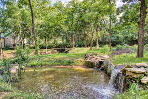 A serene pond surrounded by lush greenery and trees, with a small waterfall cascading over rocks.