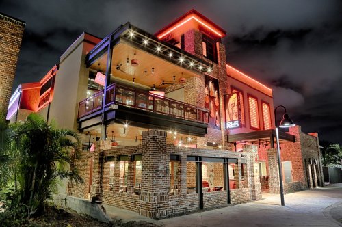 A modern restaurant exterior at night, featuring brick walls, vibrant neon lights, and a spacious balcony.