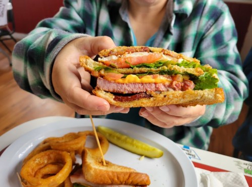 A person holds a layered sandwich with lettuce, tomato, cheese, and bacon, alongside onion rings and a pickle.