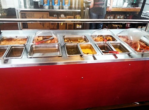 A buffet-style food station with various dishes in metal trays, covered with clear lids, on a red counter.